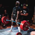 A man lifting a bar with weights on both the left side of the bar, standing on a mat that's also in the foreground, and a man standing behind him, spotting him, with people to the left, center and right in the background.