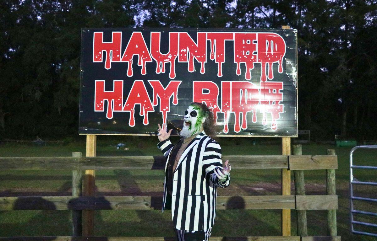 An actor dressed as the character, Beetlejuice, poses in front of a sign reading, "Haunted Hay Ride" in red, dripping font posted on a wooden fence.