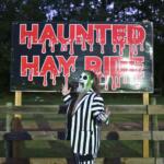 An actor dressed as the character, Beetlejuice, poses in front of a sign reading, "Haunted Hay Ride" in red, dripping font posted on a wooden fence.