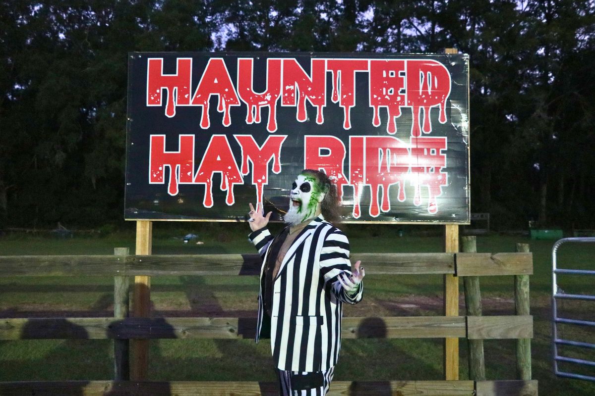 An actor dressed as the character, Beetlejuice, poses in front of a sign reading, "Haunted Hay Ride" in red, dripping font posted on a wooden fence.