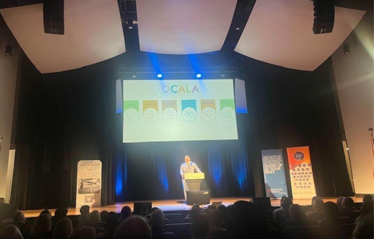 Extreme left, a flag against the wall, one poster to the left of the speaker, two posters to the right, a large screen in the background, a speaker at the podium with a microphone, in the foreground, the back of people's heads sitting in the audience.