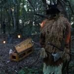 A forest has lights strewn along a path. A scarecrow with a scythe and coffin decoration stand along the path creating an eerie atmosphere.