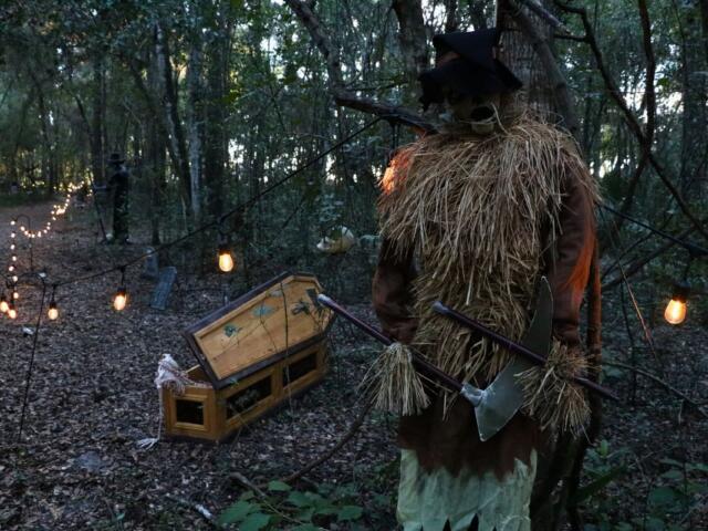 A forest has lights strewn along a path. A scarecrow with a scythe and coffin decoration stand along the path creating an eerie atmosphere.