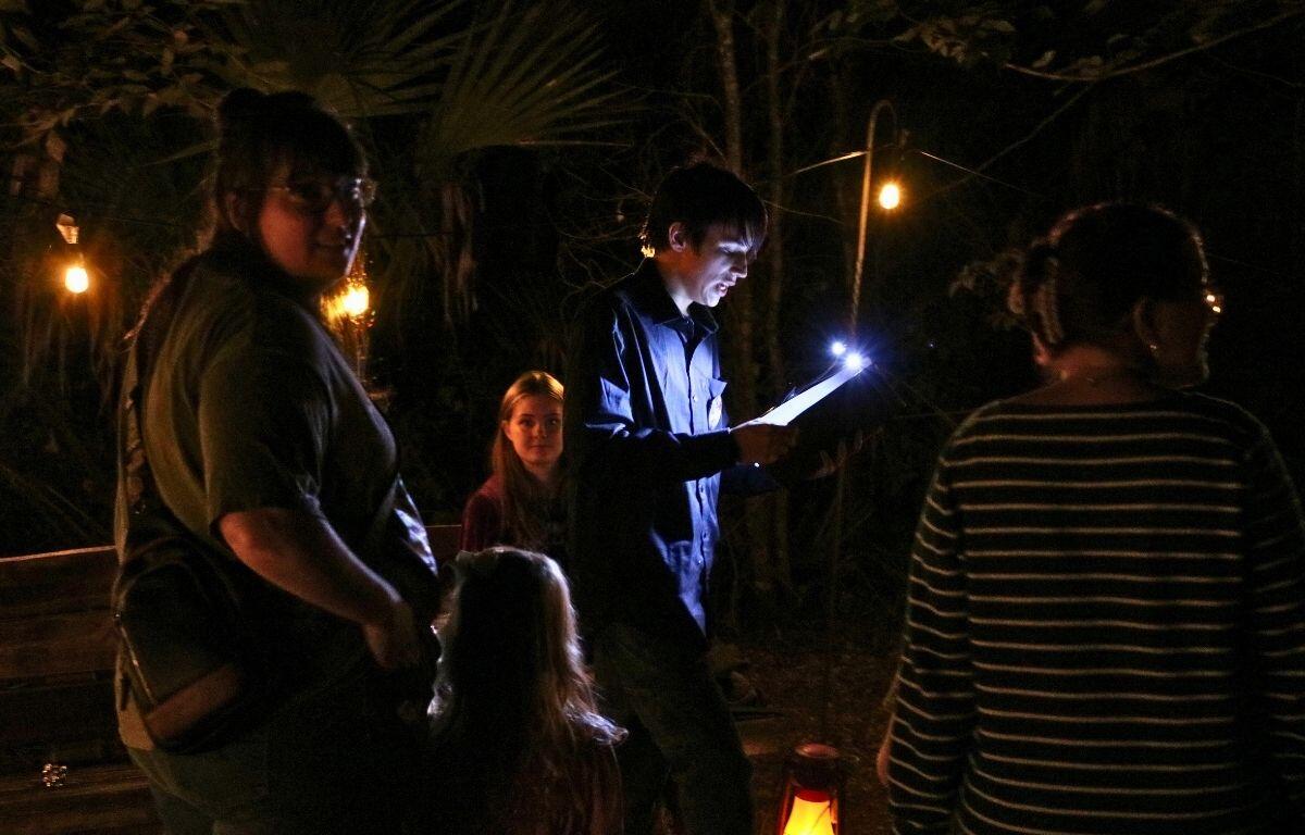 A small crowd of people stand around a man telling a story with a book in hand. The area is surrounded by a dense forest and lights strewn around. The man holds a book with a small light on top.