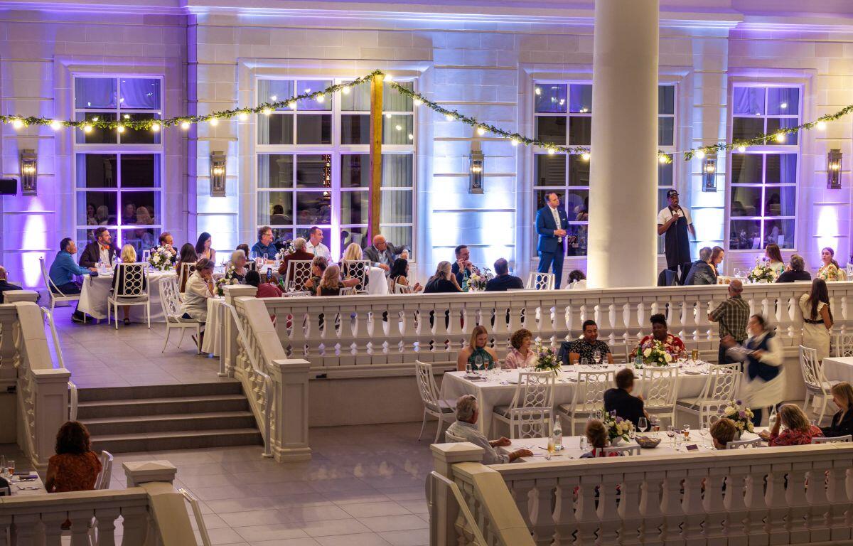 Guests seated at round tables in a spacious, well-lit hall with white decor and soft purple lighting.