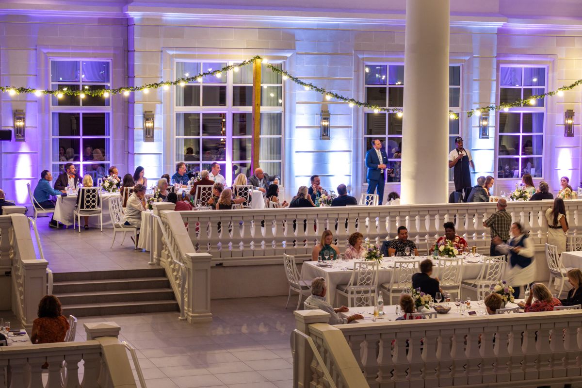 Guests seated at round tables in a spacious, well-lit hall with white decor and soft purple lighting.