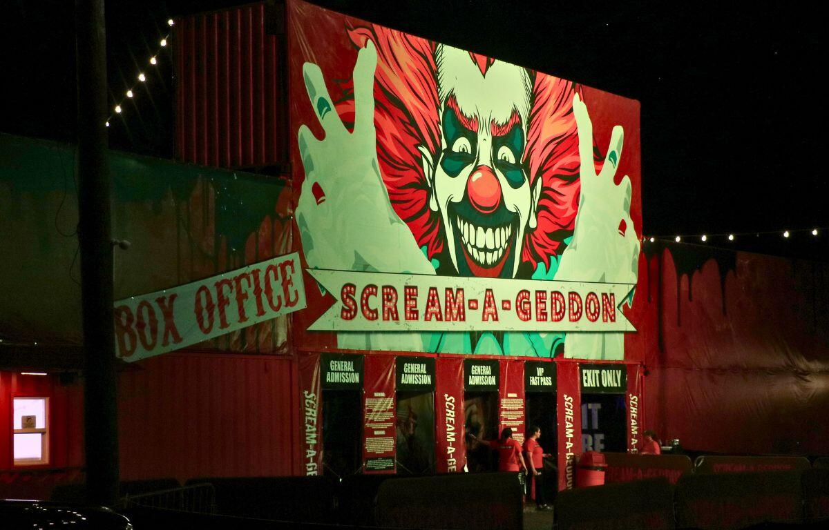 A haunted attraction has a red facade with a large clown clown painted on it. The facade has words reading, directions, "Box Office" and "Scream a Geddon."