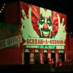 A haunted attraction has a red facade with a large clown clown painted on it. The facade has words reading, directions, "Box Office" and "Scream a Geddon."