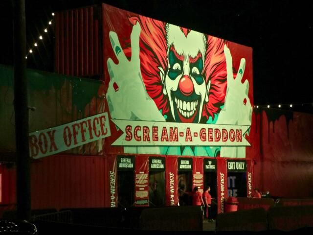 A haunted attraction has a red facade with a large clown clown painted on it. The facade has words reading, directions, "Box Office" and "Scream a Geddon."
