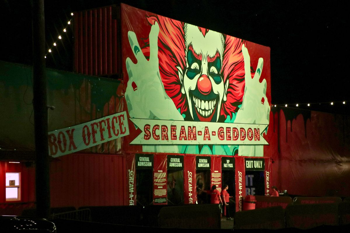 A haunted attraction has a red facade with a large clown clown painted on it. The facade has words reading, directions, "Box Office" and "Scream a Geddon."