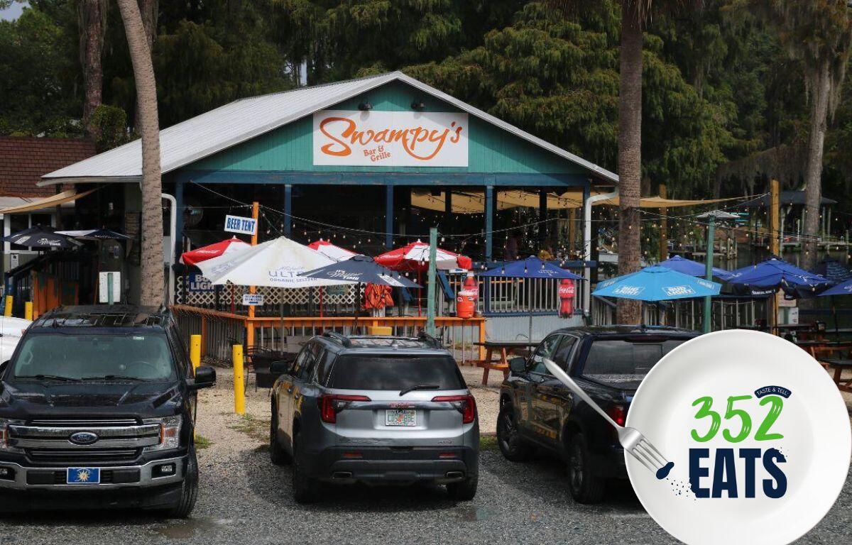 Exterior view of Swampy’s Bar & Grille with outdoor seating, colorful umbrellas, and parked vehicles in front.
