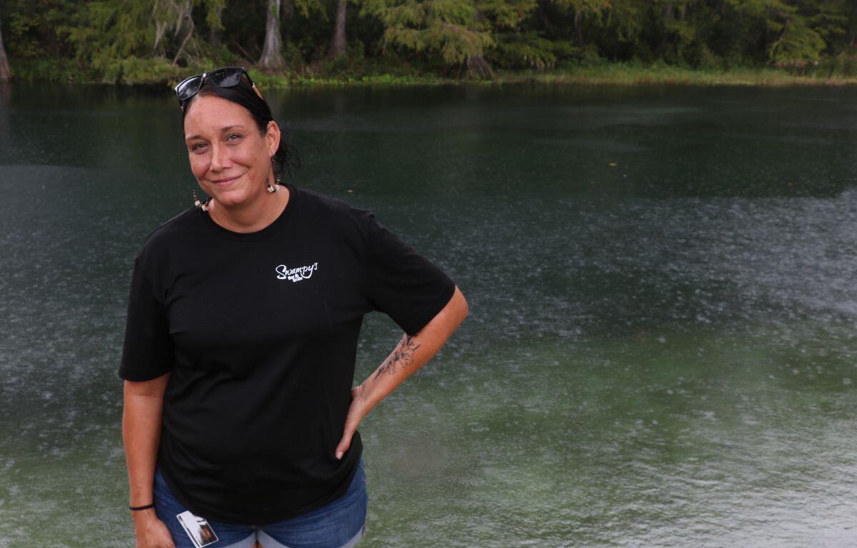 A woman wearing a Swampy’s Bar & Grille t-shirt smiles while standing beside the river.