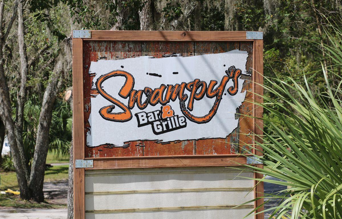 A wood sign has a white and orange logo that reads, "Swampy's Bar & Grille." in the background are trees, bushes, a sidewalk and palm plants.