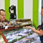 A man stands behind a display counter with a variety of chocolate items on display in the case and on top of it, a vase of purple flowers and an old-fashion phone on a green and white wall behind the man. A girl stands on the other side of the counter with a chocolate truffle in her hand.