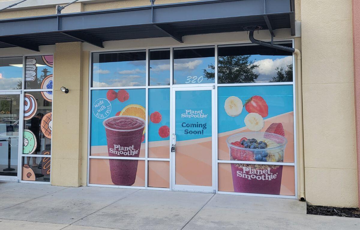 A tan colored building in a outdoor shopping plaza. Window clings are advertising a new Planet Smoothie.