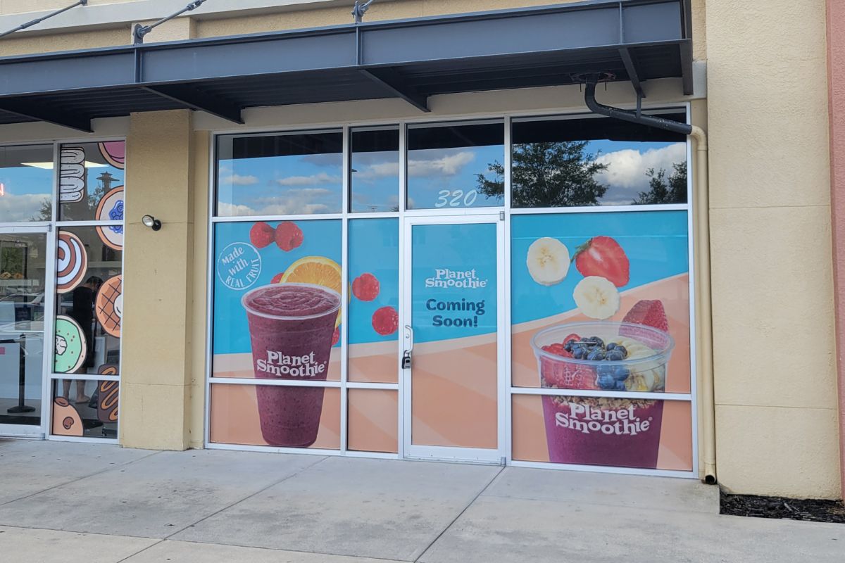A tan colored building in a outdoor shopping plaza. Window clings are advertising a new Planet Smoothie.