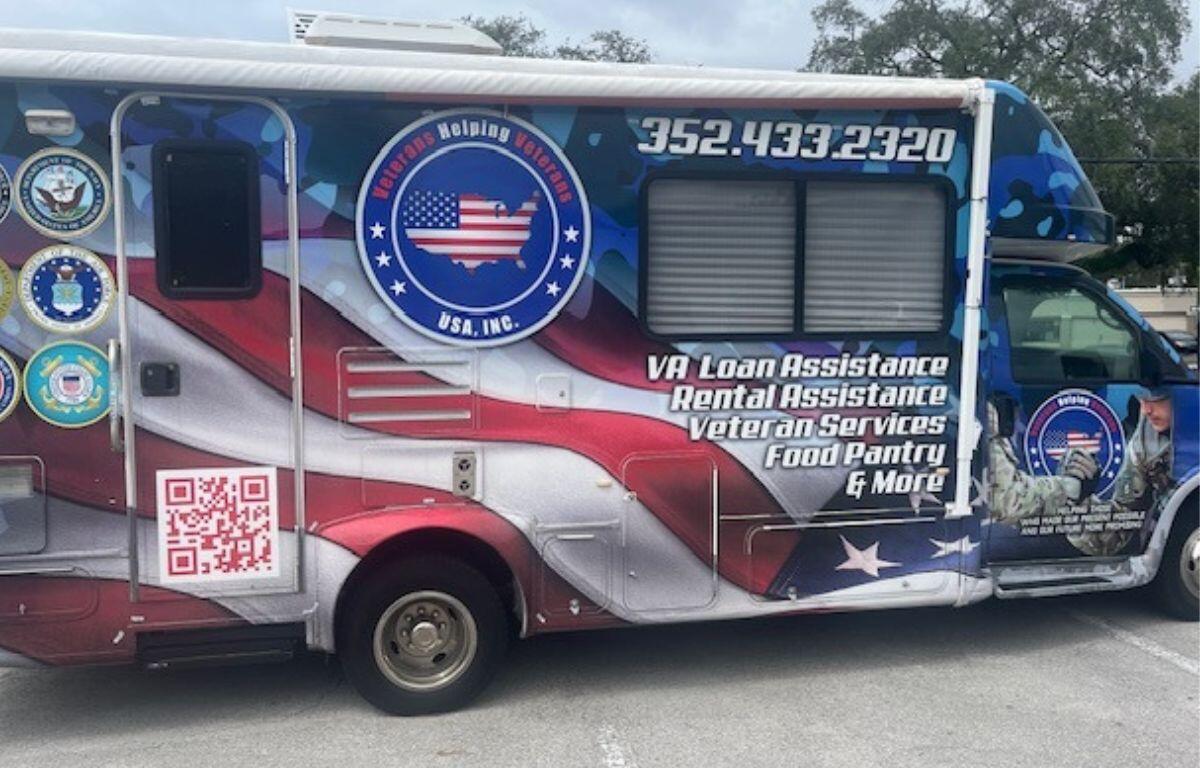 An RV with a colorful wrap, with a paved parking are in the foreground, the sky and the top of a tree line in the background.