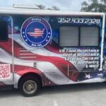 An RV with a colorful wrap, with a paved parking are in the foreground, the sky and the top of a tree line in the background.