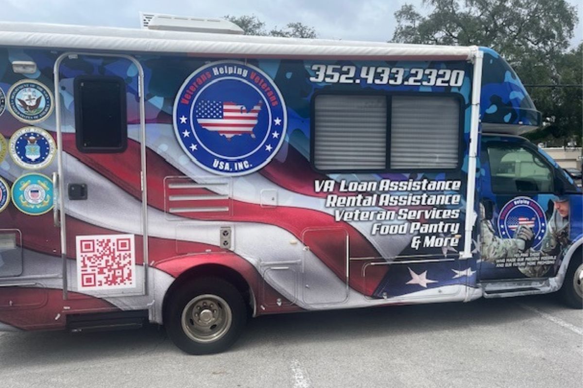 An RV with a colorful wrap, with a paved parking are in the foreground, the sky and the top of a tree line in the background.