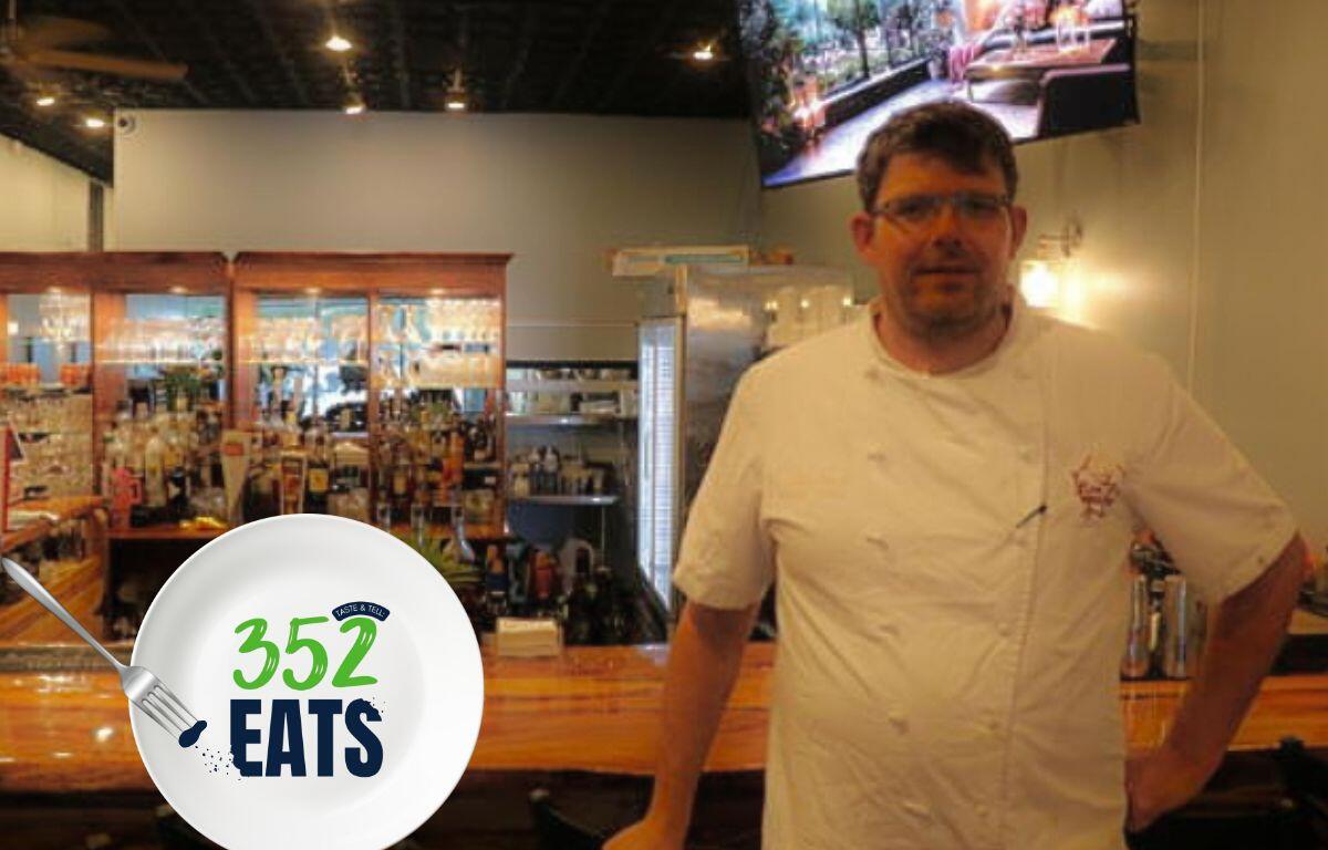 A man in a chef's outfit stands in front of a bar with a TV screen above him in the background. The bar behind him is full of glasses on wooden shelves and wooded surfaces around it.