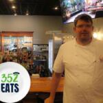 A man in a chef's outfit stands in front of a bar with a TV screen above him in the background. The bar behind him is full of glasses on wooden shelves and wooded surfaces around it.