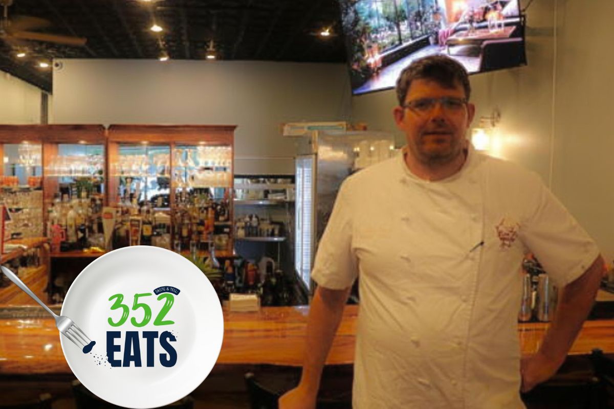 A man in a chef's outfit stands in front of a bar with a TV screen above him in the background. The bar behind him is full of glasses on wooden shelves and wooded surfaces around it.