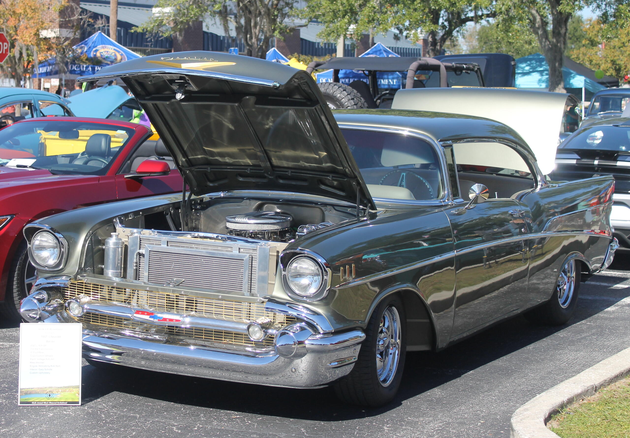 A placard to the left and a vehicle with its hood up., pavement in the foreground and to the left of the car, with cars and trees in the background.