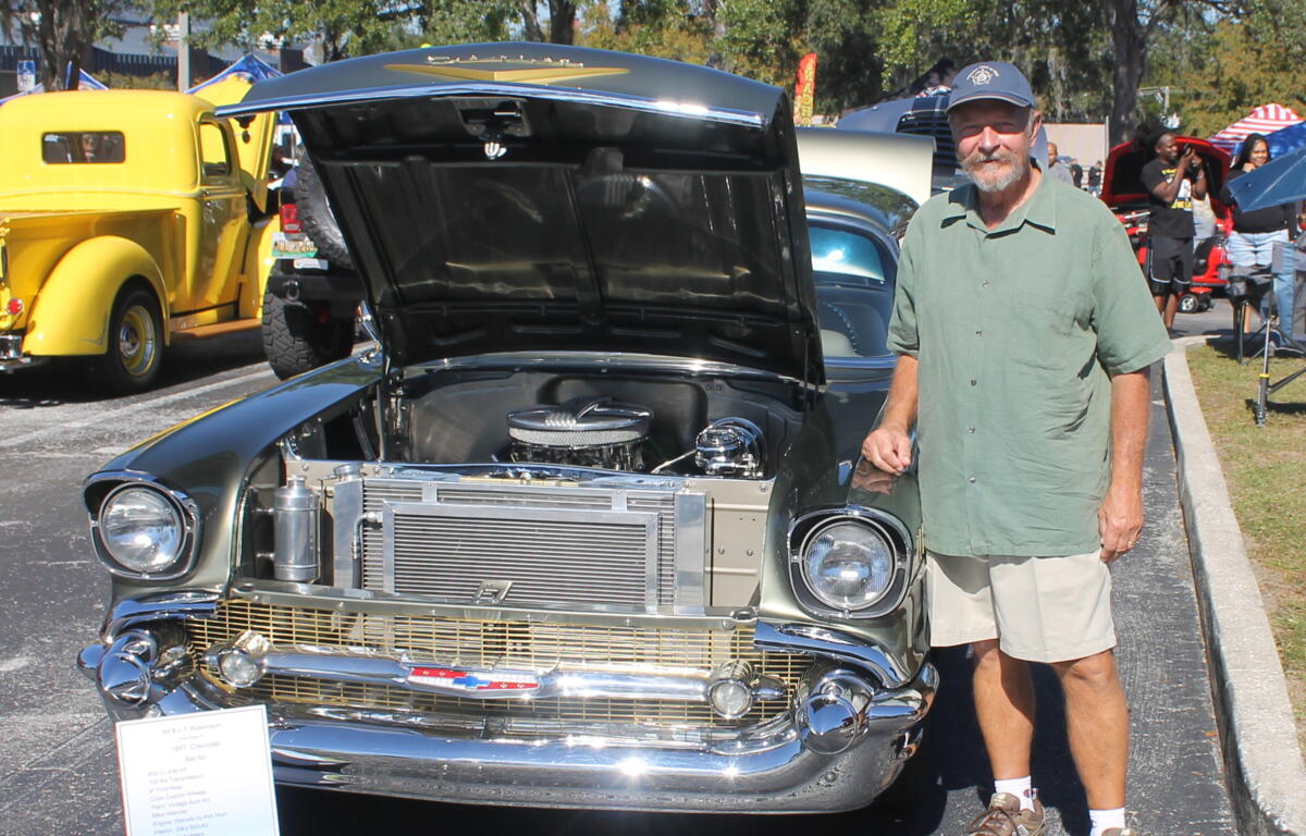 A placard to the left of the vintage classic car, with a yellow truck in the background to the left, pavement in the foreground, and a man wearing a hat, a green shirt and shorts to the right of the vehicle.