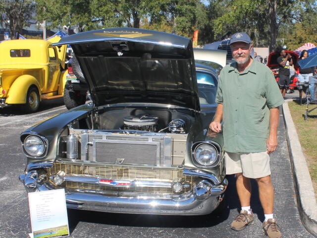 A placard to the left of the vintage classic car, with a yellow truck in the background to the left, pavement in the foreground, and a man wearing a hat, a green shirt and shorts to the right of the vehicle.