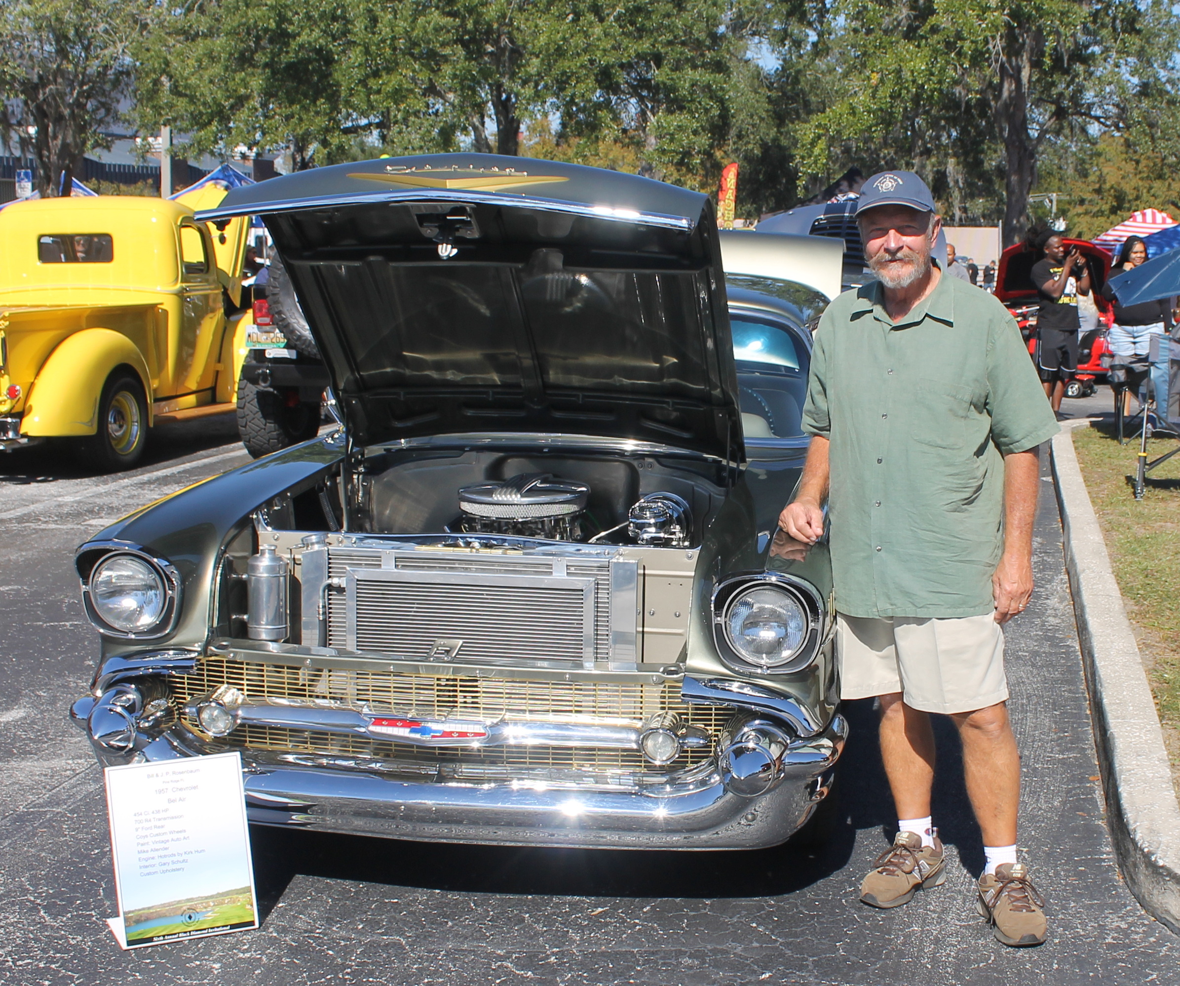 A placard to the left of the vintage classic car, with a yellow truck in the background to the left, pavement in the foreground, and a man wearing a hat, a green shirt and shorts to the right of the vehicle.