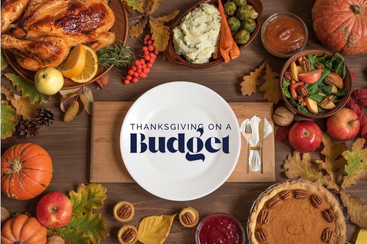 A Thanksgiving table filled with budget-friendly holiday foods, including a roasted chicken, mashed potatoes, vegetables, salad, cranberries, pie, pumpkins and apples, with a large white plate in the center displaying the text “Thanksgiving on a Budget.”
