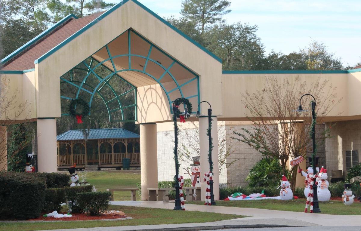 An open pavilion with lamp posts along a paved walkway leading toward it has Christmas decorations around it. The decorations include candy canes, wreaths snowman and deflated inflatable characters.