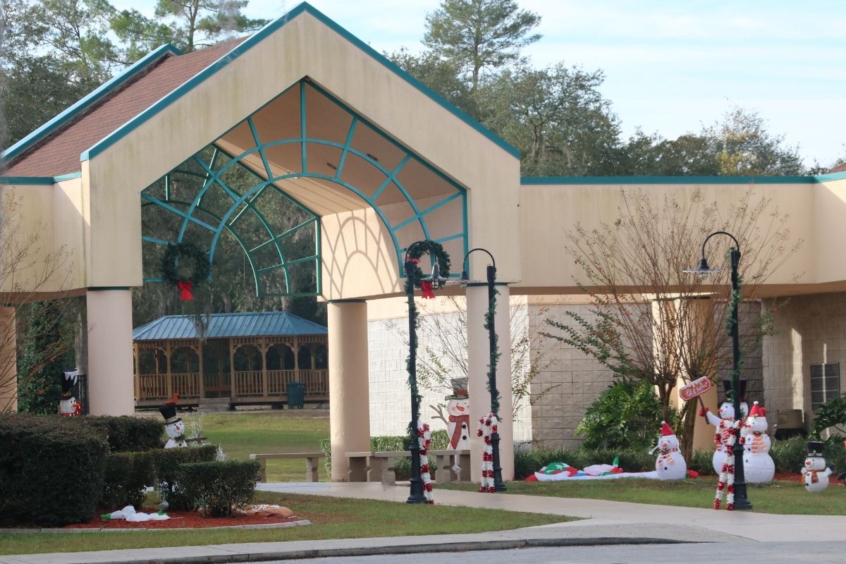 An open pavilion with lamp posts along a paved walkway leading toward it has Christmas decorations around it. The decorations include candy canes, wreaths snowman and deflated inflatable characters.