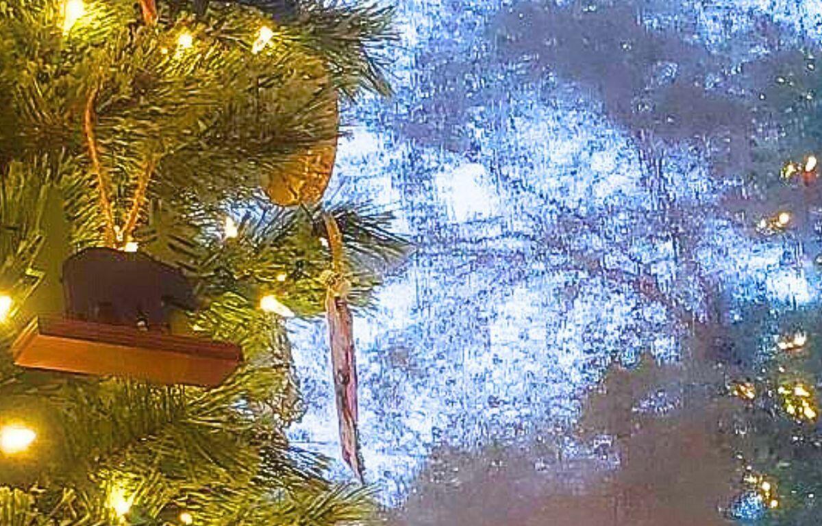 A Christmas tree with bright string lights, a bear ornament and other decorations is reflected in a window with rain falling against it.