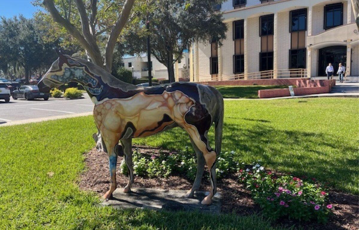A fiberglass horse, stabilized over grass, with trees, two men talking and a building in the background.