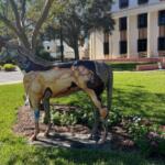 A fiberglass horse, stabilized over grass, with trees, two men talking and a building in the background.