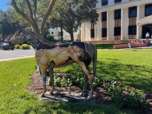 A fiberglass horse, stabilized over grass, with trees, two men talking and a building in the background.