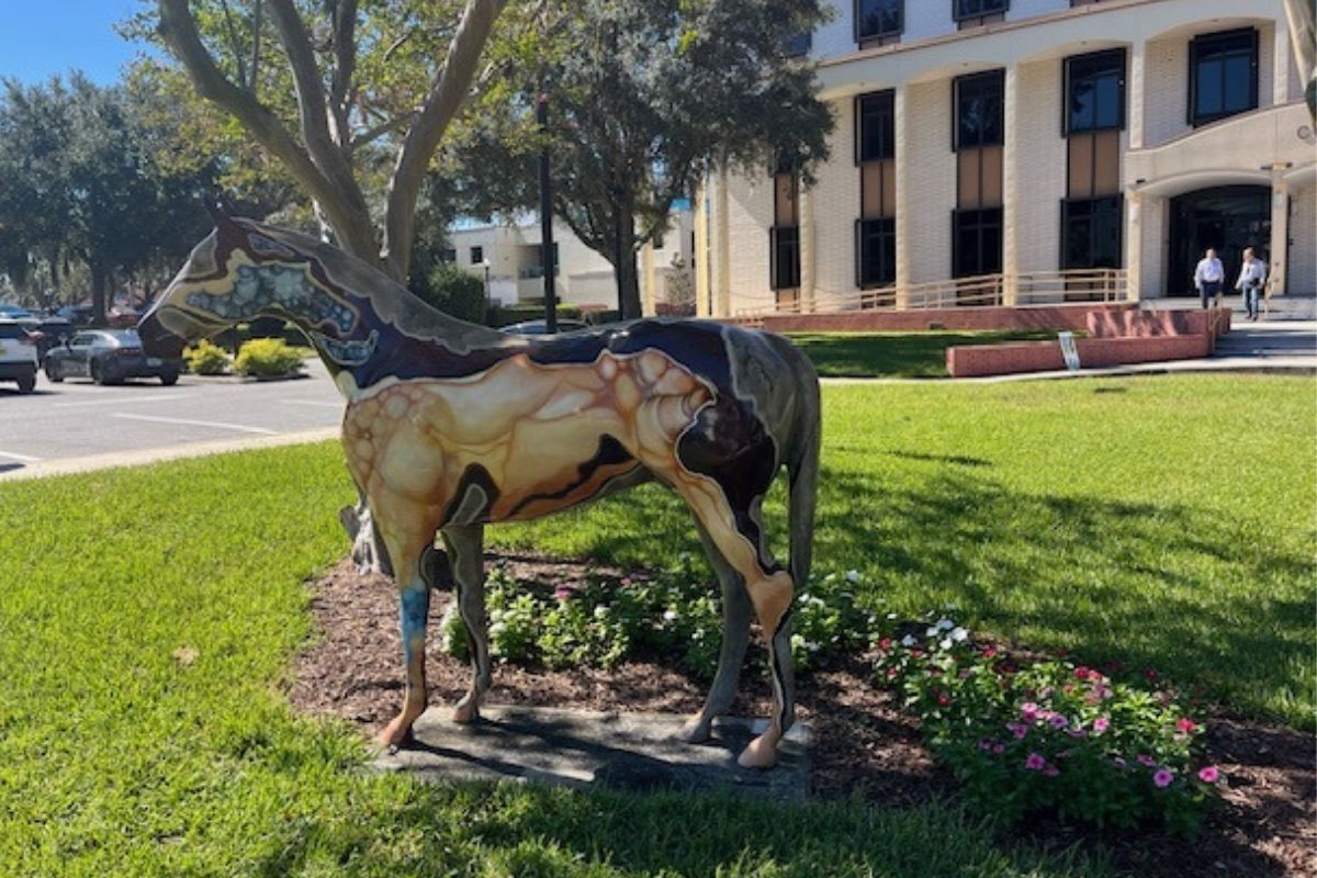 A fiberglass horse, stabilized over grass, with trees, two men talking and a building in the background.