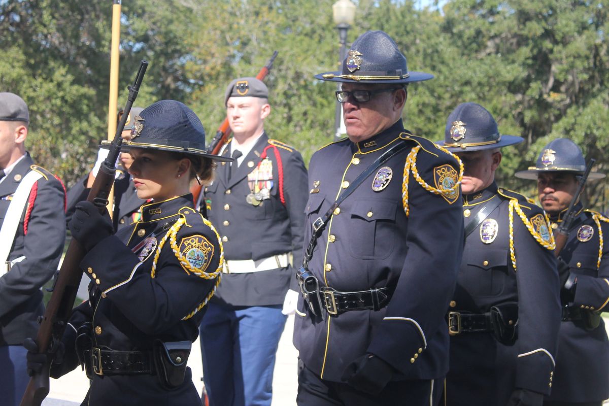 A series of people dressed in uniform in both the foreground and background, with a woman on the left with a rifle, people in the background carrying rifles and flags.