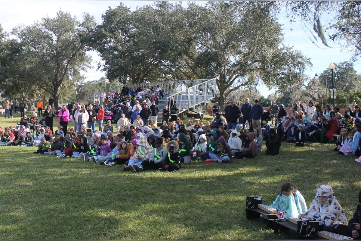 Grass in the foreground, with two kids wrapped in blankets, with a slew of people dressed in winter coats standing in front of bleacher risers, that are in front of trees and a brick wall, with a field of grass in the middle ground of the image.