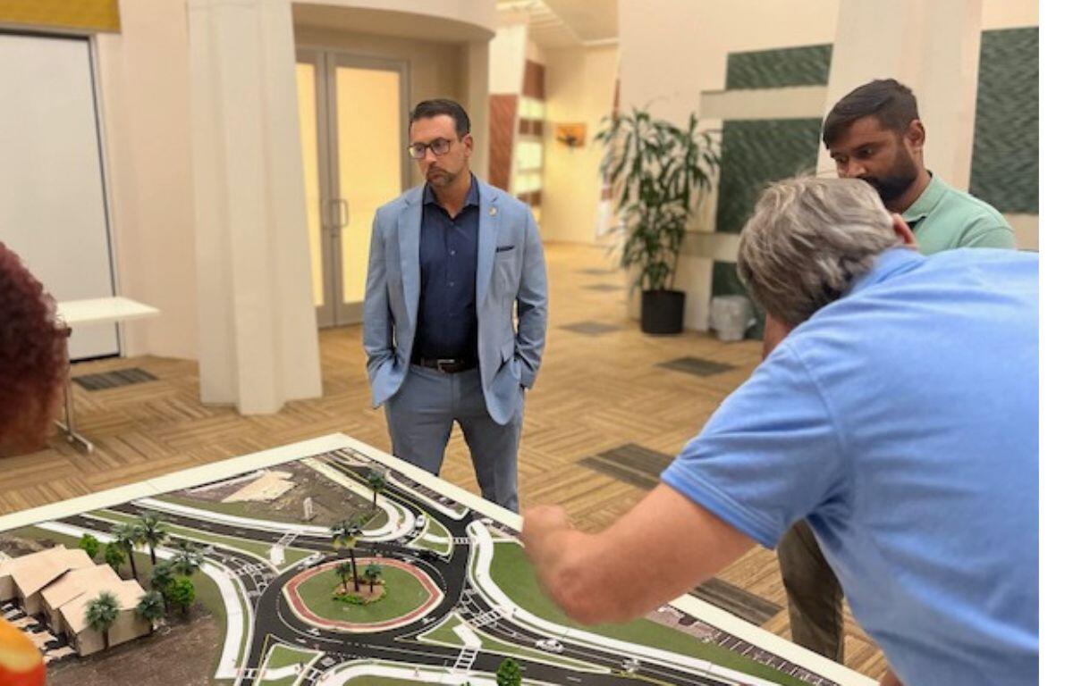 A board with a scale model on a table with someone to the left, and two people to the right in the foreground, with a man looking ponderously at the model, with doors, plants and windows in the background.