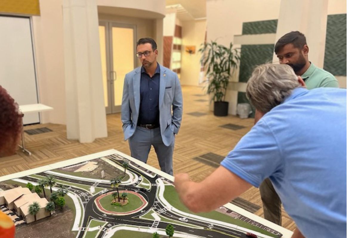 A board with a scale model on a table with someone to the left, and two people to the right in the foreground, with a man looking ponderously at the model, with doors, plants and windows in the background.