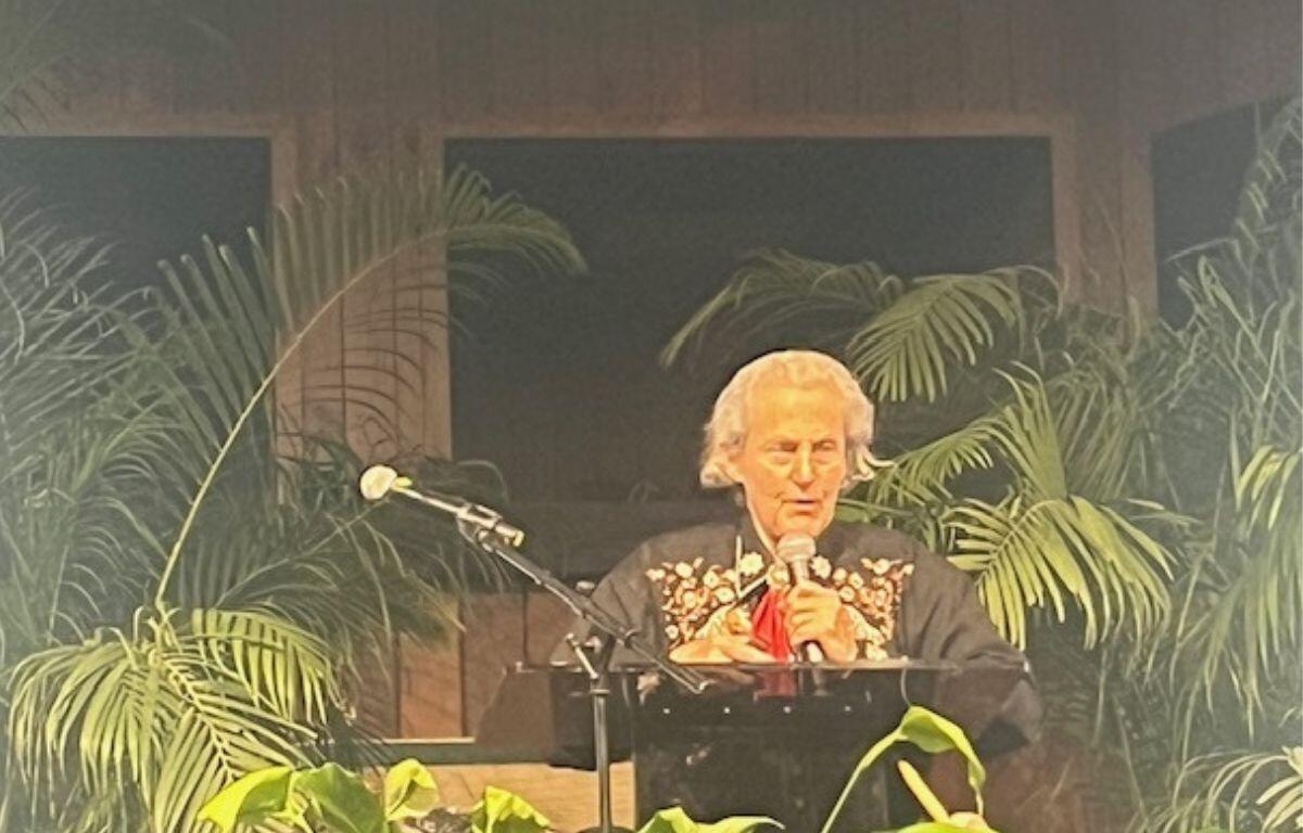 Background has wood paneling and windows, center image a woman dressed in western wear, behind a podium and microphone and plant and a microphone in the foreground.