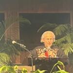 Background has wood paneling and windows, center image a woman dressed in western wear, behind a podium and microphone and plant and a microphone in the foreground.