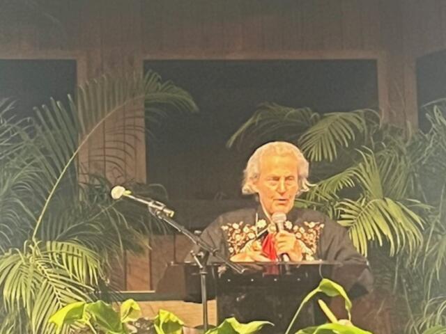 Background has wood paneling and windows, center image a woman dressed in western wear, behind a podium and microphone and plant and a microphone in the foreground.