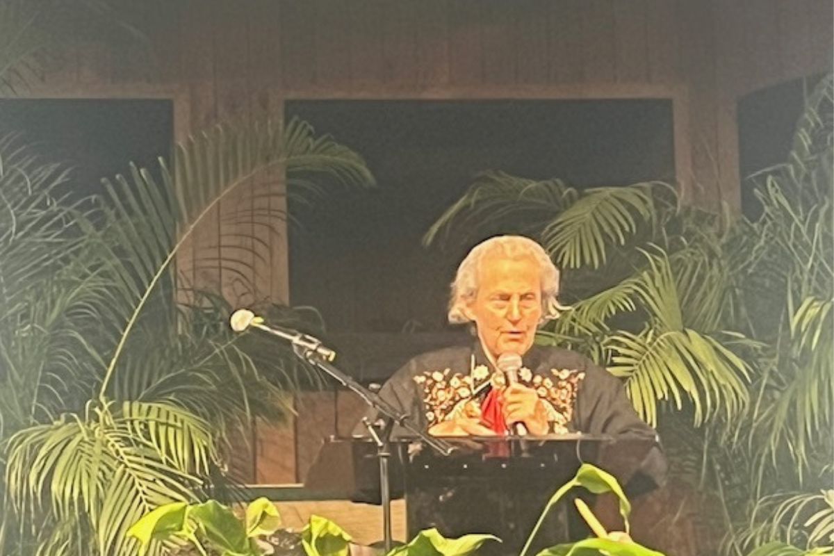 Background has wood paneling and windows, center image a woman dressed in western wear, behind a podium and microphone and plant and a microphone in the foreground.