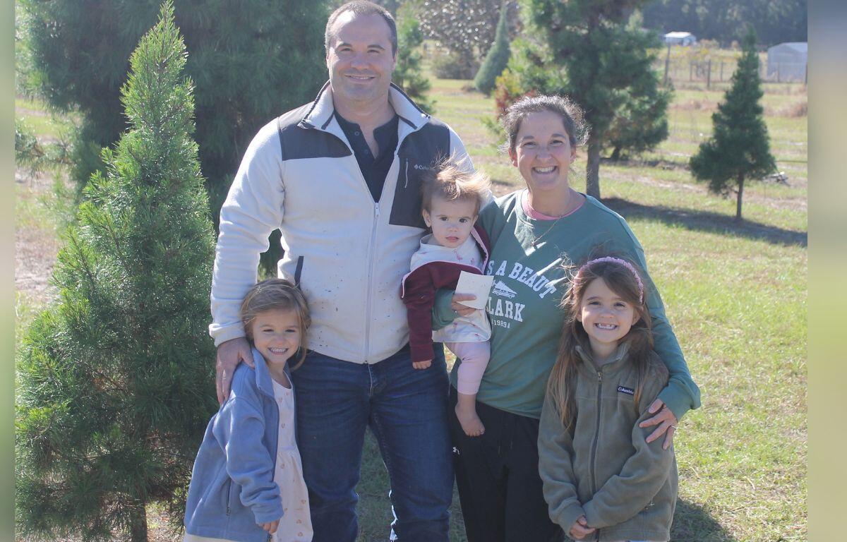 A tree to the left of a family, with one daughter to the left of the two parents who are holding a child and another daughter to the right, who are standing on grass with trees in the background.