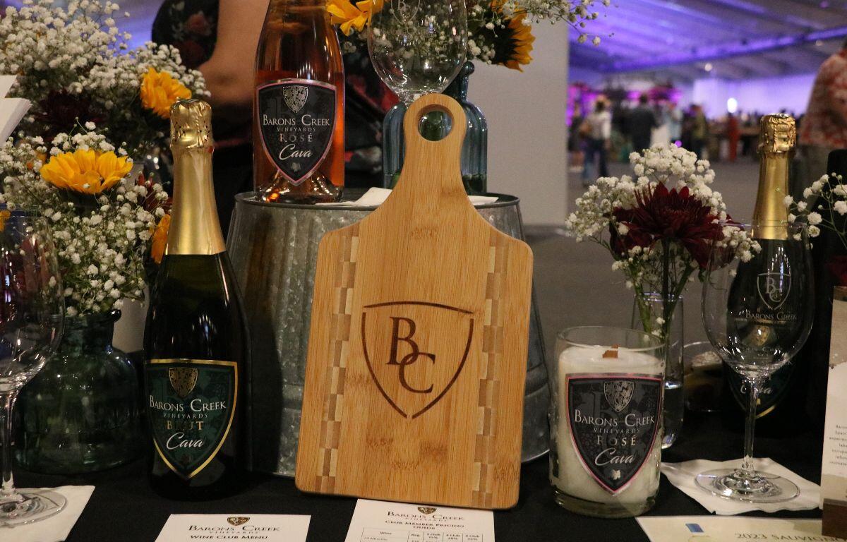 A table features a board with a logo reading, "BC." On the table is also an upside-down bucket with a bottle of alcohol, a wine glad and a vase with flowers. Around the board are more alcohol bottles, a wine glass and papers.