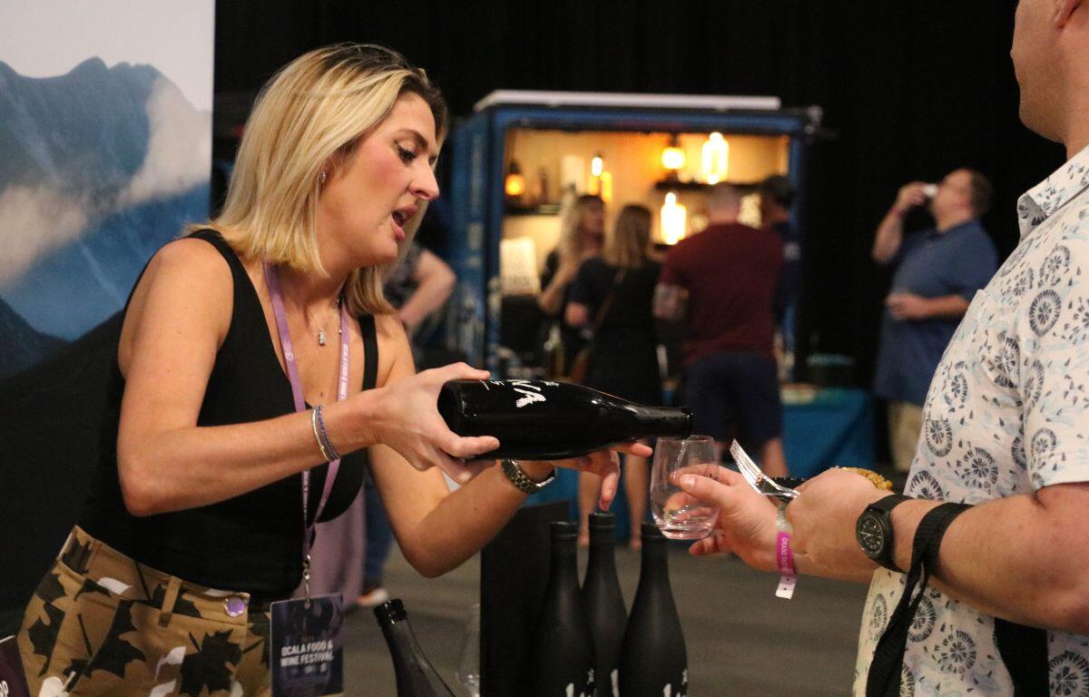 A woman pours alcohol from a bottle into a clear wine glass for a man. In the background is a booth with people lining up for more drinks.