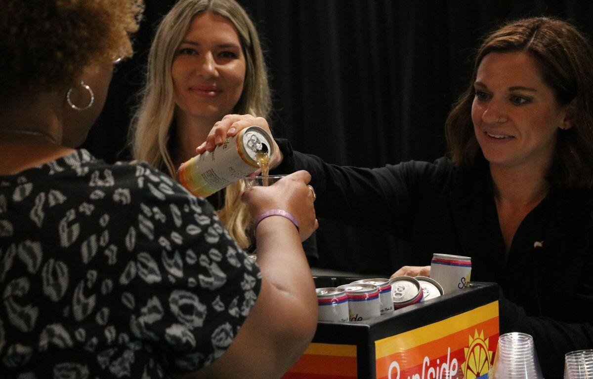 Two women stand behind a counter with container filled with Surfside branded canned drinks and clear cups next to it. One of the women pours some of the can into a cup for another woman.